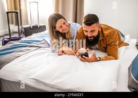 Young happy couple is on vacation. Tourists arrived in hotel room with their suitcases. They are lying on bed and using smartphone. Stock Photo