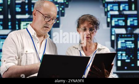 Dilligent manager and assistant looking around data center, using laptop and tablet to crosscheck disaster recovery plan and assess high tech workspace units in need of replacement, preventing risks Stock Photo