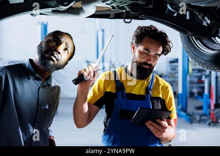 Technician in garage using tablet to follow checklist while doing maintenance on car, talking with client. Employee in auto repair shop does checkup on vehicle tires next to customer using lamp light Stock Photo