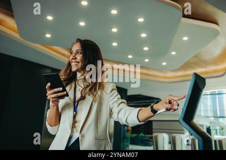 A confident professional woman interacts with a touchscreen device in a sleek, modern corporate office. She is smiling, holding a smartphone, and enga Stock Photo