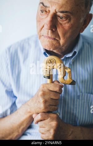 An elderly man sits quietly, holding a beautifully carved cane. His thoughtful demeanor reflects a mix of nostalgia and contemplation in a serene indo Stock Photo
