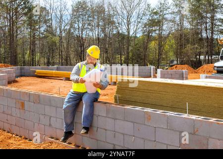 Construction worker in helmet sits on concrete block wall, examining plans on construction site surrounded by trees building materials under bright sunlight. Stock Photo