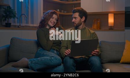 Caucasian male using smartphone on couch home evening loving wife hugging husband smiling man and woman customers shopping in online store on Stock Photo