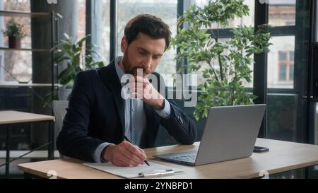Pensive puzzled Caucasian business man businessman male entrepreneur corporate employee sales manager working in office typing on laptop searching Stock Photo