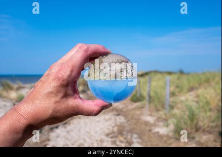 A human hand holds a glass ball, in the ball a sand dune, a footpath, blue sky and the sea are reflected Stock Photo