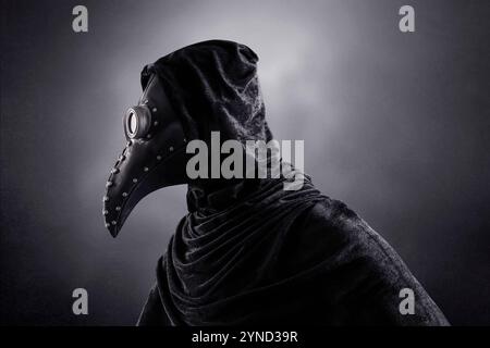 Plague doctor in hooded cloak at night over dark misty background Stock Photo