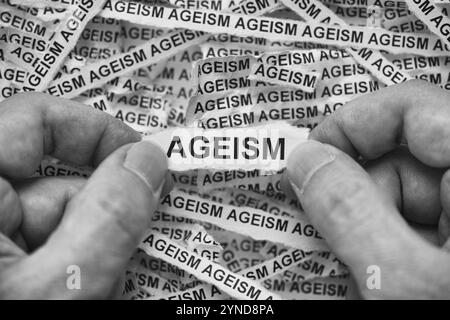 A man holding a piece of paper with the word Ageism on it in his hands over crumpled strips of newspaper. Black and White. Close up. Stock Photo