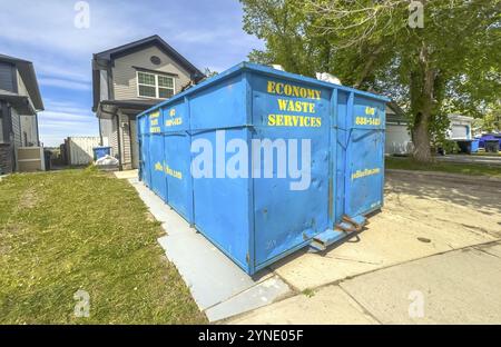 Calgary, Alberta, Canada. Jun 19, 2024. A blue metal dumpster sits on a driveway in front of a house with a white roof. There is a yellow sign with te Stock Photo
