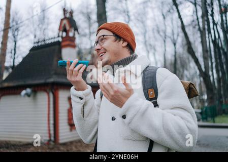 Young man with backpack using voice assistant on his smartphone while exploring a city park, enjoying travel and technology Stock Photo