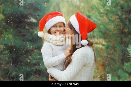 Family, celebration, holiday. Happy mother kissing her little girl child in santa hat together near Christmas tree in forest with snow Stock Photo