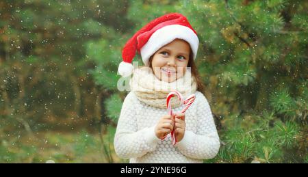 Christmas portrait of happy smiling little girl child in santa red hat with sweet lollipop cane stick outdoors on green tree background with snow Stock Photo