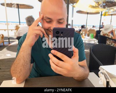 Smiling young man using his cell phone in a beach restaurant on a sunny day, looking at the mobile phone laughing Stock Photo
