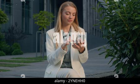 Caucasian woman girl female businesswoman business lady checking email message using phone outdoors reading good news on smartphone showing thumbs up Stock Photo
