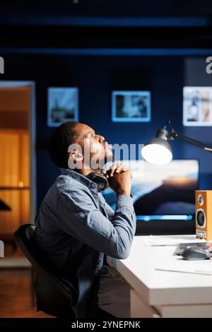 Tired african american guy experiences burnout as he works from home, struggling to handle corporate duties using global web analytics. At desk, independent contractor worker is worn out. Stock Photo