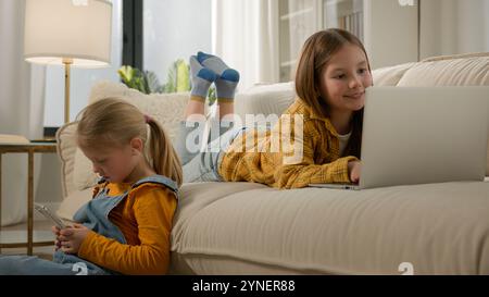Two Caucasian kids girls sisters daughters friends using different gadgets in living room play online games together on laptop mobile phone watching Stock Photo