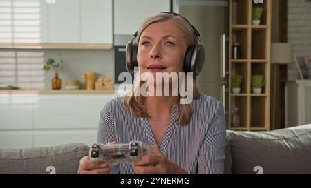 Funny smiling mature caucasian woman playing video game console in headphones using joystick controller at home on couch elderly female play computer Stock Photo