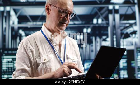 Aged professional navigating network of server units in industrial hub. Technician with laptop doing inspection and optimizations in high tech data center, preventing system difficulties Stock Photo