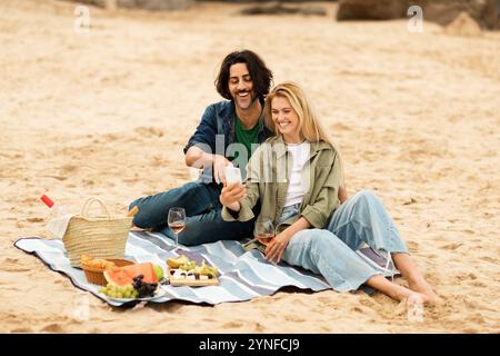 Couple enjoying a picnic on the beach while taking a selfie together Stock Photo