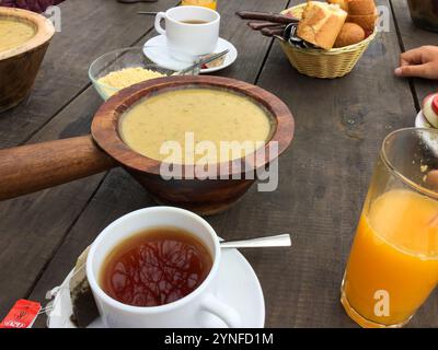 A rich traditional dish is presented in a wooden bowl, accompanied by beverages including tea and orange juice. Nearby, a basket holds bread rolls, co Stock Photo