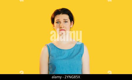 Melancholy disappointed woman in blue dress standing studio portrait isolated on yellow empty space background. Stock Photo
