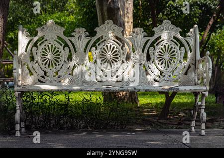 One empty bench made of white iron stands on city park. Sunlight on backrest surface of Victorian pattern white cast iron garden bench in a shady area Stock Photo