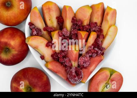 Whole, cut in half and sliced kissabel apple sprinkled with frozen raspberries served in heart shaped bowl against white background. Hidden rose apple Stock Photo