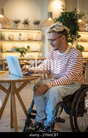 Smiling disabled man in wheelchair works on laptop at cafe table. Freelancer working remotely online Stock Photo