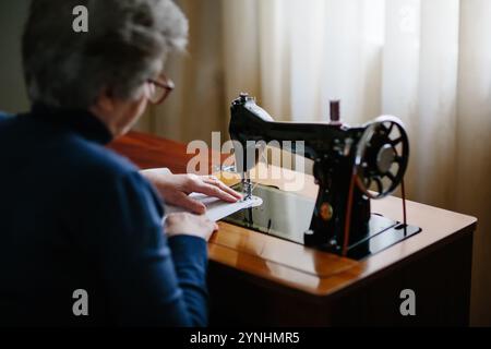 A senior woman focuses on sewing fabric on a classic sewing machine in a cozy home environment. Soft natural light filters through curtains, enhancing Stock Photo