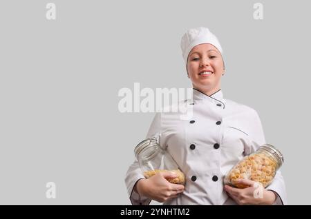Female chef in white uniform holding two glass jars filled with different types of pasta, smiling at camera on gray background Stock Photo