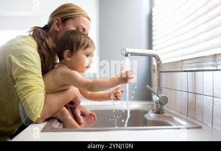 A mother and her young son engage in playful exploration at the kitchen sink. The boy reaches for the running water, showing his curiosity, while his Stock Photo