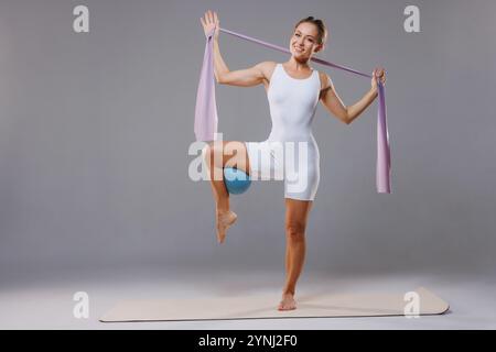 Portrait of sporty adult woman in white bodysuit, holding pilates and fitness rubber band and blue ball, stretching workout Stock Photo
