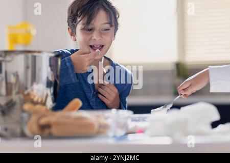 A young boy smiles while tasting a dessert in a well-lit kitchen. Another hand prepares nearby ingredients, creating a cheerful cooking moment. Stock Photo