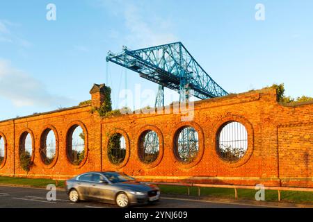 car speeds past old red brick wall vulcan salt works with circular openings & disused blue transporter bridge in background middlesbrough Teesside uk Stock Photo