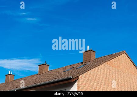 A residential building's brown tiled roof, featuring three chimneys, is set against a vibrant blue sky with a few wispy clouds. Stock Photo