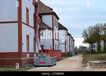 A residential construction site displays a large debris chute extending from a building to a dumpster below, with a gravel pathway nearby and overcast Stock Photo