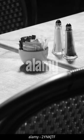 Sugar bowl with brown and white sugar sachets, salt cellar and pepper caster on marble table at an outdoor cafe. Wicker chairs. Selective focus on the Stock Photo