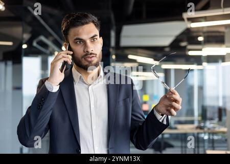 Businessman taking important phone call in office. Holds glasses while conversing, conveys confidence, communication, professionalism. Modern business Stock Photo