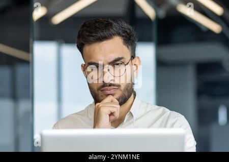 Man wearing glasses concentrating on computer screen in office setting. Serious expression suggests deep thought or analysis. Modern work environment. Keywords: office, focus, professional. Stock Photo