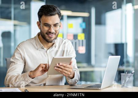 Smiling young professional using tablet in modern office. Male office worker multitasking, combining digital devices like tablet and laptop for efficient work and communication. Stock Photo