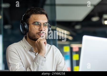 Focused young man in modern office wearing headphones, engaged in thoughtful online work session on laptop screen, embodying concentration, innovation, and technology-driven workspace. Stock Photo