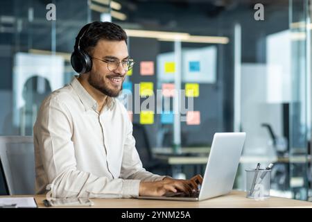 Smiling man in headphones works at laptop in modern office. Appears focused and engaged with task. Office supplies on desk add detail to professional environment. Stock Photo