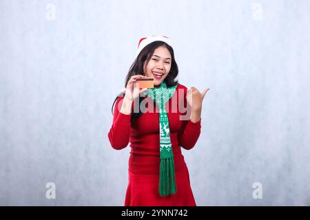 gesture of young Asian ladies wearing red christmas sweater, Santa hat and scarf, cheerful looking forward holding credit debit card and pointing left Stock Photo