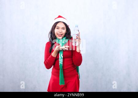 gesture of Asian ladies wearing red christmas sweater, Santa hat, and scarf, cheerful hand holding white water bottle and offering it, isolated on whi Stock Photo