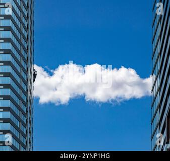 White, fluffy cloud between two modern high rise buildings, urban architecture and nature. A window cleaning platform on the left building, Vibrant bl Stock Photo