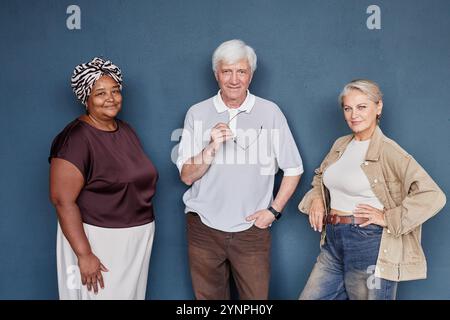 Group portrait of three senior people wearing casual clothing posing together on blue background in studio all smiling at camera Stock Photo