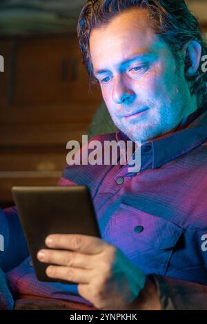 A man is sitting in a chair and smiling while looking at a tablet reading a digital book. He is enjoying the content on the screen. Stock Photo