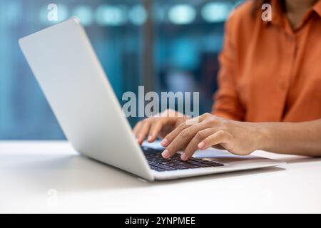 Hands typing on laptop in office. Business professional in orange shirt working efficiently. Captures modern office atmosphere, digital communication, and professional work culture. Stock Photo
