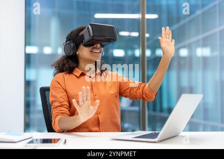 Woman using VR headset at desk, immersed in virtual environment. Professional setting highlights technology's role in innovation, creativity, and productivity in office work. Stock Photo