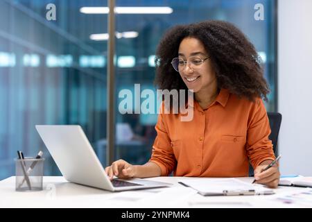 Confident office worker in modern workspace using laptop and taking notes. She embodies productivity and joy in professional environment, showcasing corporate lifestyle and digital communication. Stock Photo
