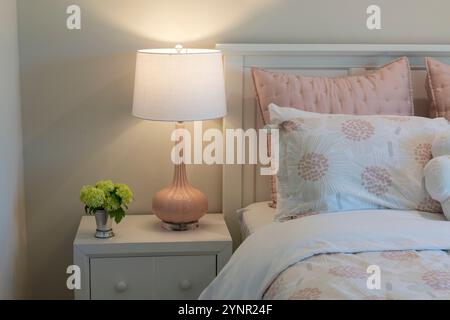 Delightful kids bedroom corner featuring pastel bedding, a pink bedside lamp, and a white nightstand with a floral touch, evoking warmth and charm. Stock Photo